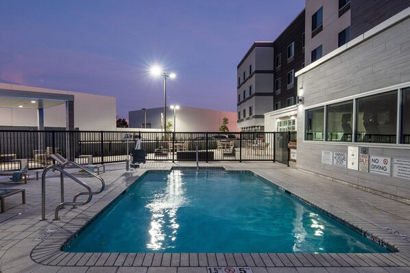 A rectangular pool with a metal ladder and a fence, surrounded by a paved area with benches and a building with large windows.