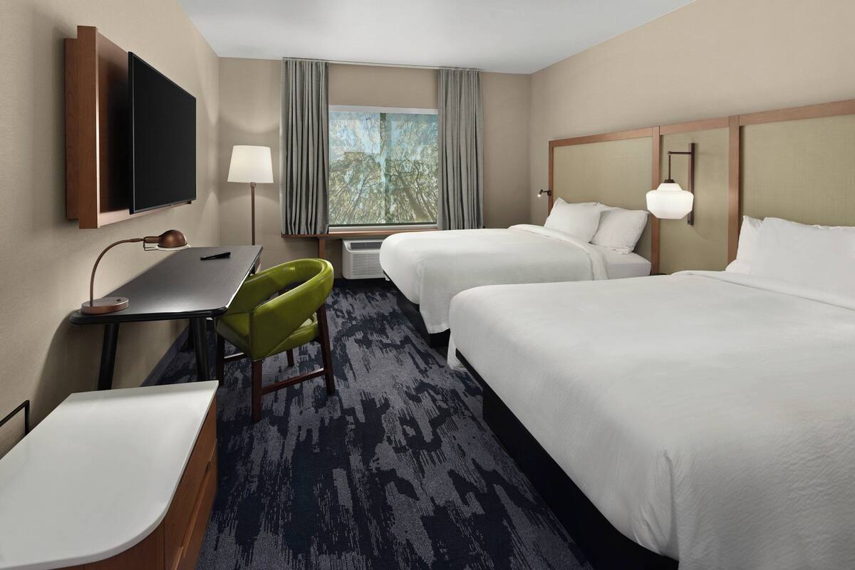 A hotel room with two beds, a desk with a chair, a TV, and a window with curtains.