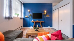 A room with a blue accent wall, a mounted TV, a wooden table, and a patterned floor. - Downtown Penthouse #1 | Vintage 2bd (+Gym) (Chicago)