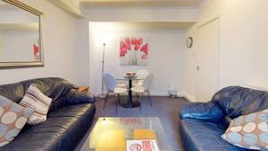 Een woonkamer met twee zwarte leren banken, een glazen salontafel, en een eethoek met een kleine ronde tafel en twee stoelen.