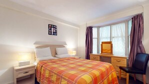Hotelkamer met een bed, nachtkastje, lamp, bureau, stoel en een raam met gordijnen.
