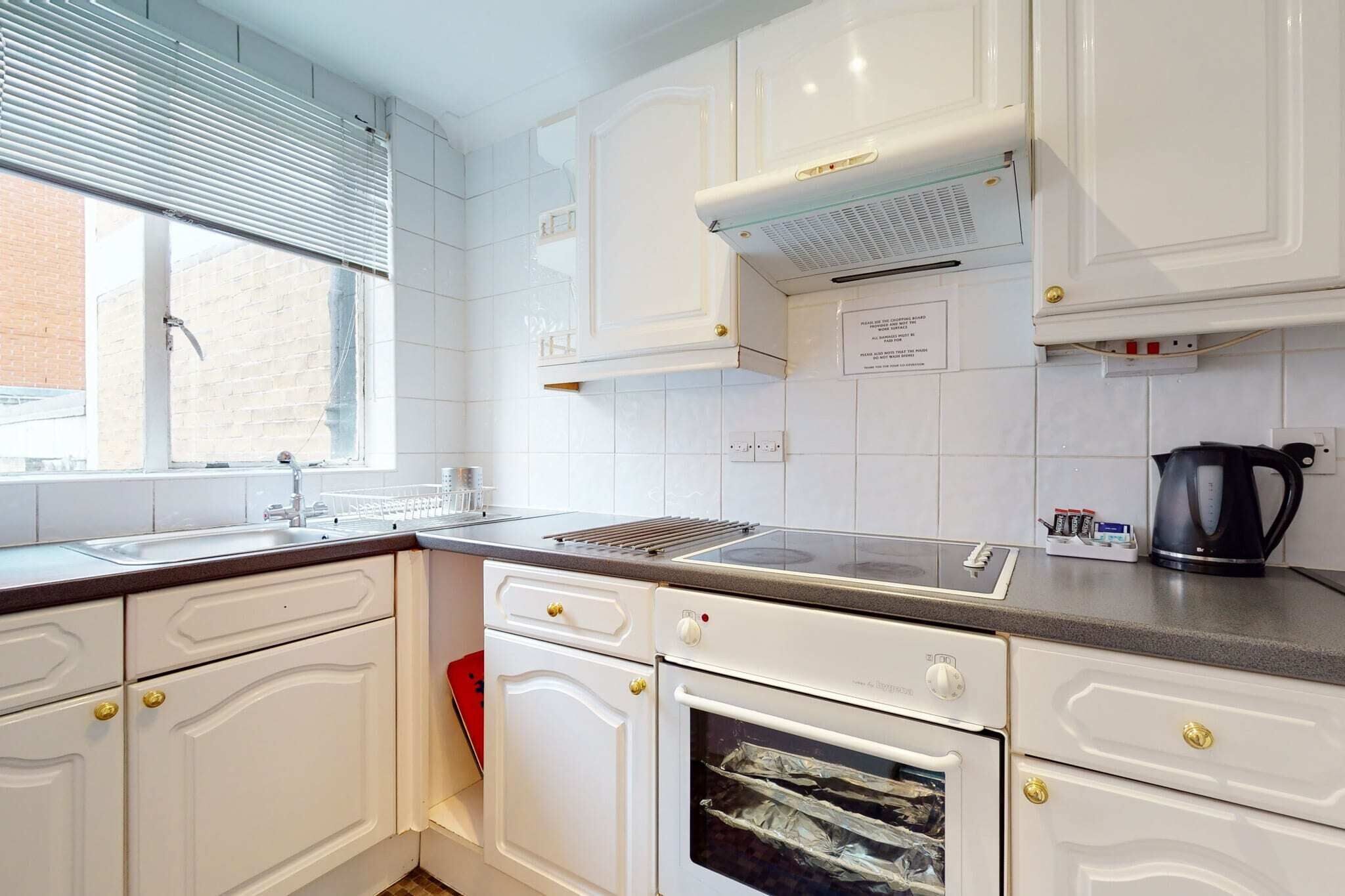 Een keuken met witte kasten, een inbouwoven, een kookplaat en een raam met jaloezieën.