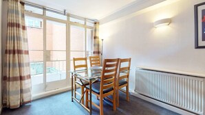 A dining area with a glass table and wooden chairs, a radiator, a window with curtains, and a wall-mounted light fixture. - Luxury Serviced Apartment, Staffed Reception, Two Bathrooms, Large Living Room a (London)