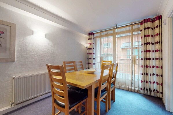 A dining area with a wooden table and chairs, a white radiator, and a window with patterned curtains.