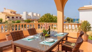 A dining table set for four with wicker chairs, a glass top, and a view of buildings and greenery.