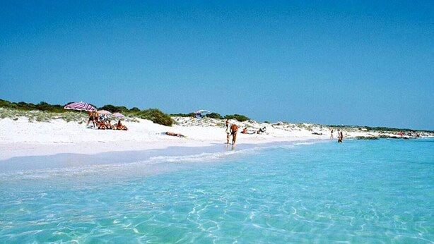 A beach with clear turquoise water, white sand, and people walking along the shore.