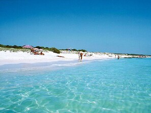 Ein Strand mit klarem türkisblauem Wasser, weißem Sand und Menschen, die am Ufer entlanggehen.