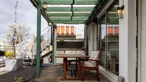 Eine Holzdielen-Terrasse mit Tisch und Stühlen, einer hängenden Pflanze und Blick auf Gebäude durch eine Glastür.