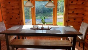 Interior de una cabaña de madera con una mesa de comedor, dos sillas, un banco y una ventana con vista a un bosque.