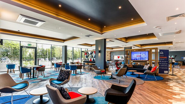 A modern hotel lobby with a variety of seating options, a large TV screen, and a view of the outdoors. - Holiday Inn Express Bicester by IHG (Bicester)