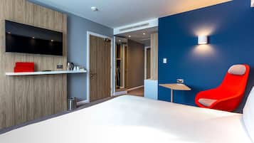 A modern hotel room with a large bed, a red chair, a flat-screen TV mounted on a wooden wall, and a small table with a lamp.
