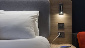 A modern hotel room with a neatly made bed, a bedside table with a smartphone charging, and a wall-mounted light fixture.