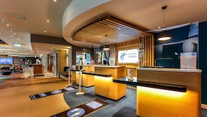 A modern hotel reception area with a wooden reception desk, a digital screen displaying the hotel name, and directional floor markers. - Holiday Inn Express Bicester by IHG (Bicester)