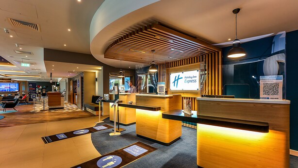 A modern hotel reception area with a wooden reception desk, a digital screen displaying the hotel name, and directional floor markers. - Holiday Inn Express Bicester by IHG (Bicester)