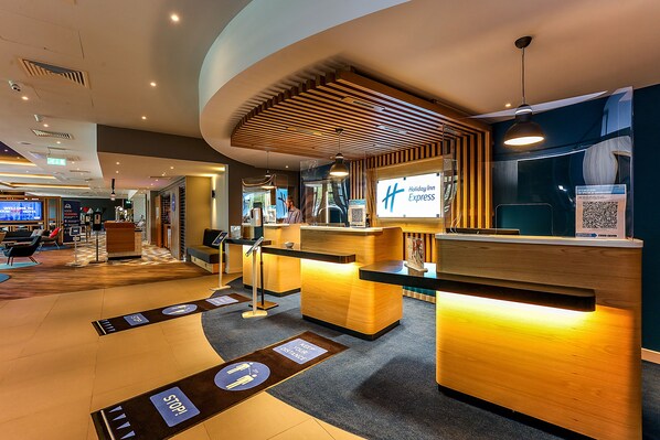 A modern hotel reception area with a wooden reception desk, a digital screen displaying the hotel name, and directional floor markers. - Holiday Inn Express Bicester by IHG (Bicester)