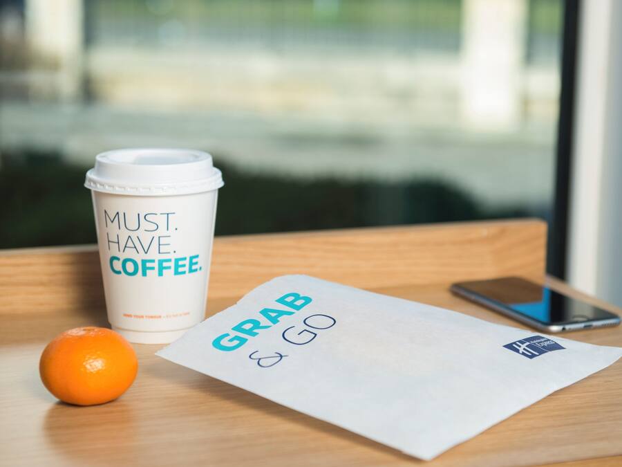 A white coffee cup with text, a piece of paper with 'GRAB & GO' written on it, a smartphone, and an orange on a wooden surface.