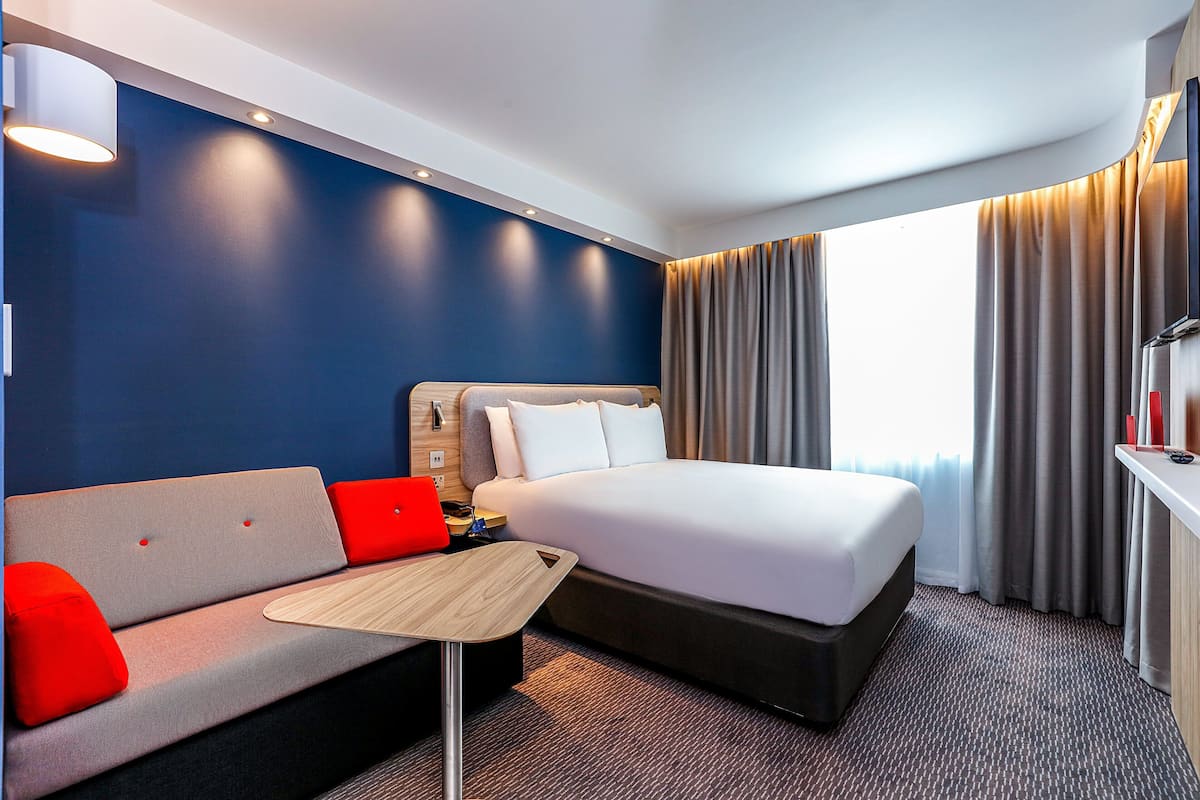 A hotel room with a bed, a sofa with red cushions, a small table, and a TV on a stand.