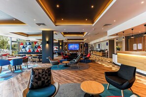 Un moderno vestíbulo de hotel con mostrador de recepción, zona de estar con sillas y mesas, y una pantalla grande mostrando un mensaje de bienvenida.