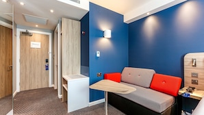 A modern room with a blue accent wall, a grey and red sofa, a small table, and a wooden cabinet. - Holiday Inn Express Bicester by IHG (Bicester)