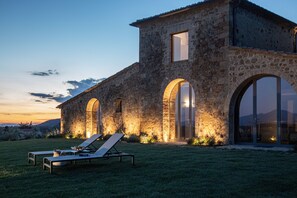A stone building with large arched windows and a well-lit entrance. There are two lounge chairs in the foreground.