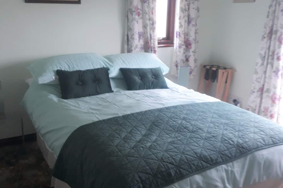 A neatly made bed with a quilted cover, a window with floral curtains, and a framed picture on the wall.