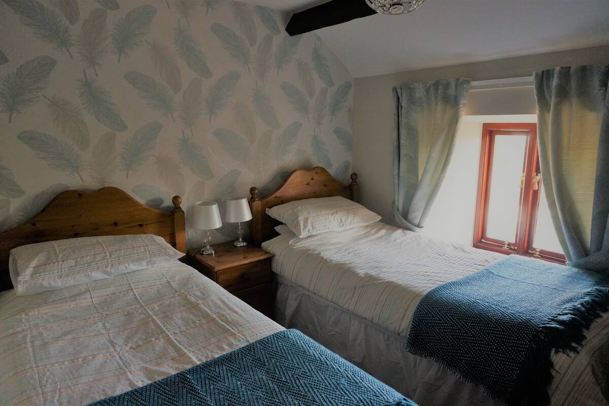 A bedroom with two beds, a nightstand with a lamp, a window with curtains, and a leaf-patterned wall.