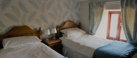 A bedroom with two beds, a nightstand with a lamp, a window with curtains, and a leaf-patterned wall.
