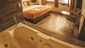 A modern hotel room with a large bathtub, a bed with an orange bedspread, a small round table, and wicker chairs. - Toucan Platinum Suites Aparthotel (Mindo)