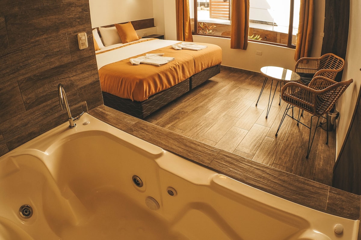 A modern hotel room with a large bathtub, a bed with an orange bedspread, a small round table, and wicker chairs.