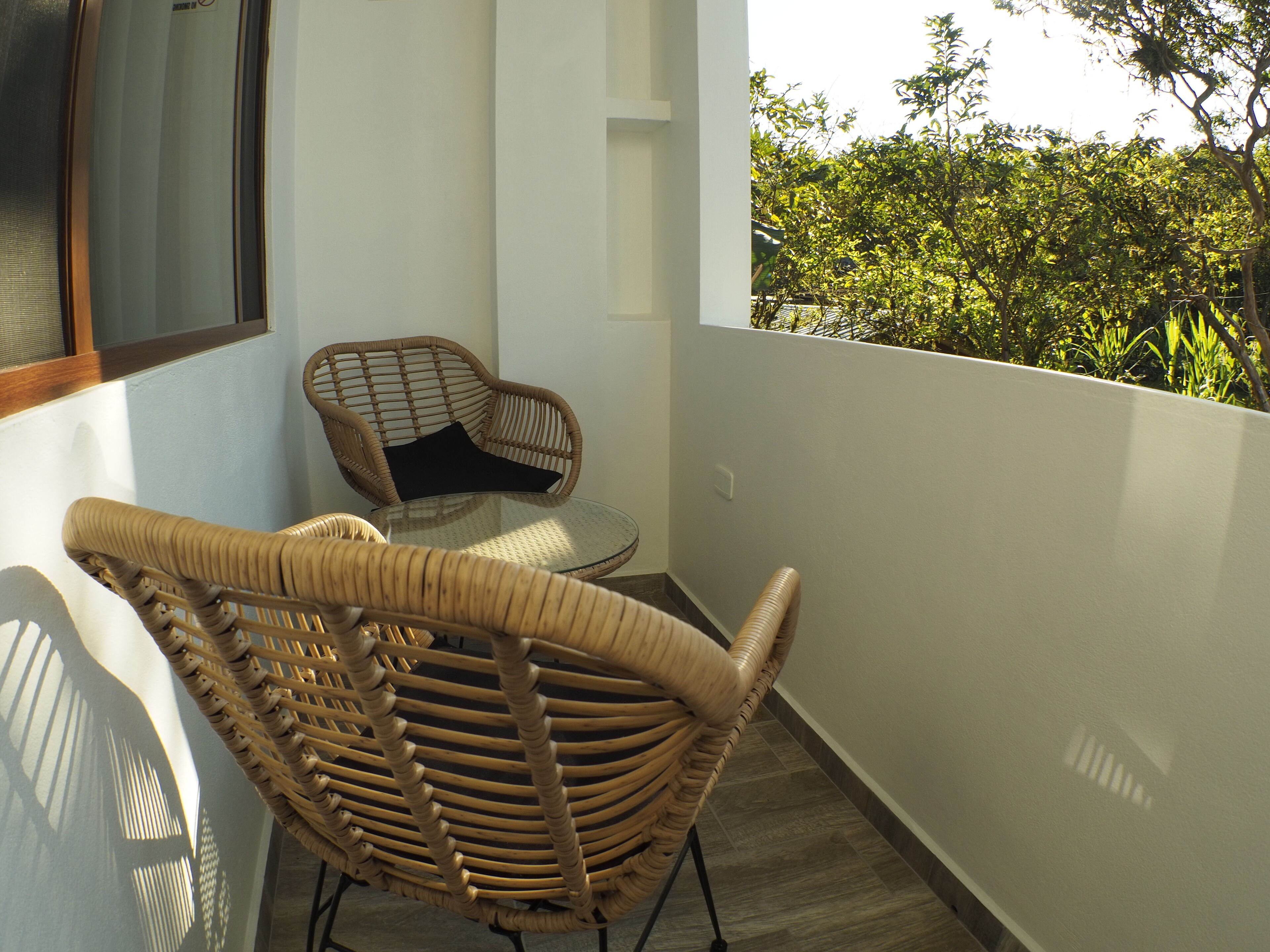 Un balcón con sillas de mimbre y una mesa de vidrio, con vista a un jardín exuberante.