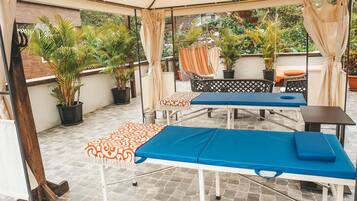 A covered outdoor area with a massage table, lounge chairs, and potted plants.