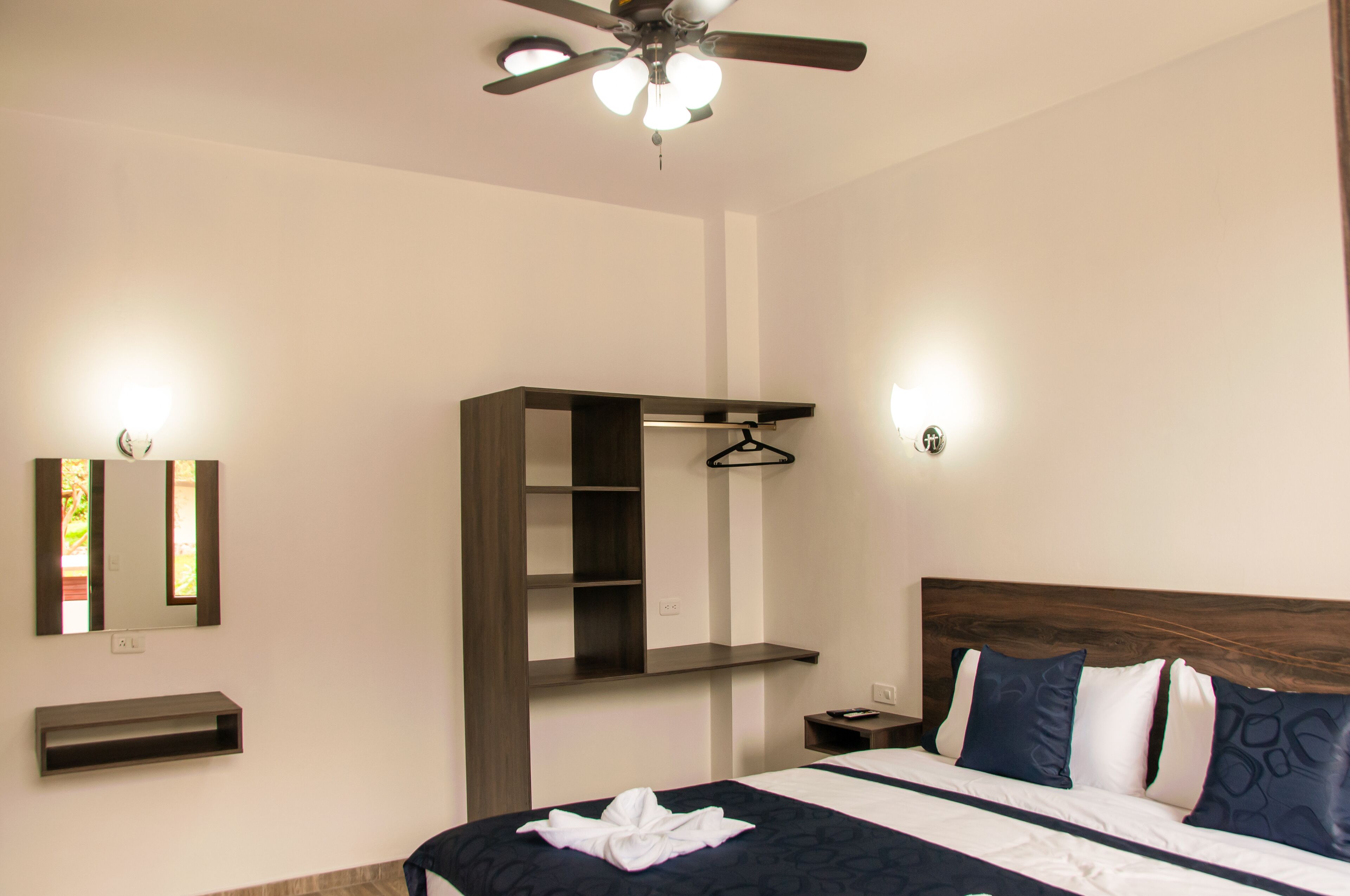 Habitación de hotel con cama, ventilador de techo, estante montado en la pared, mesita de noche y perchero.