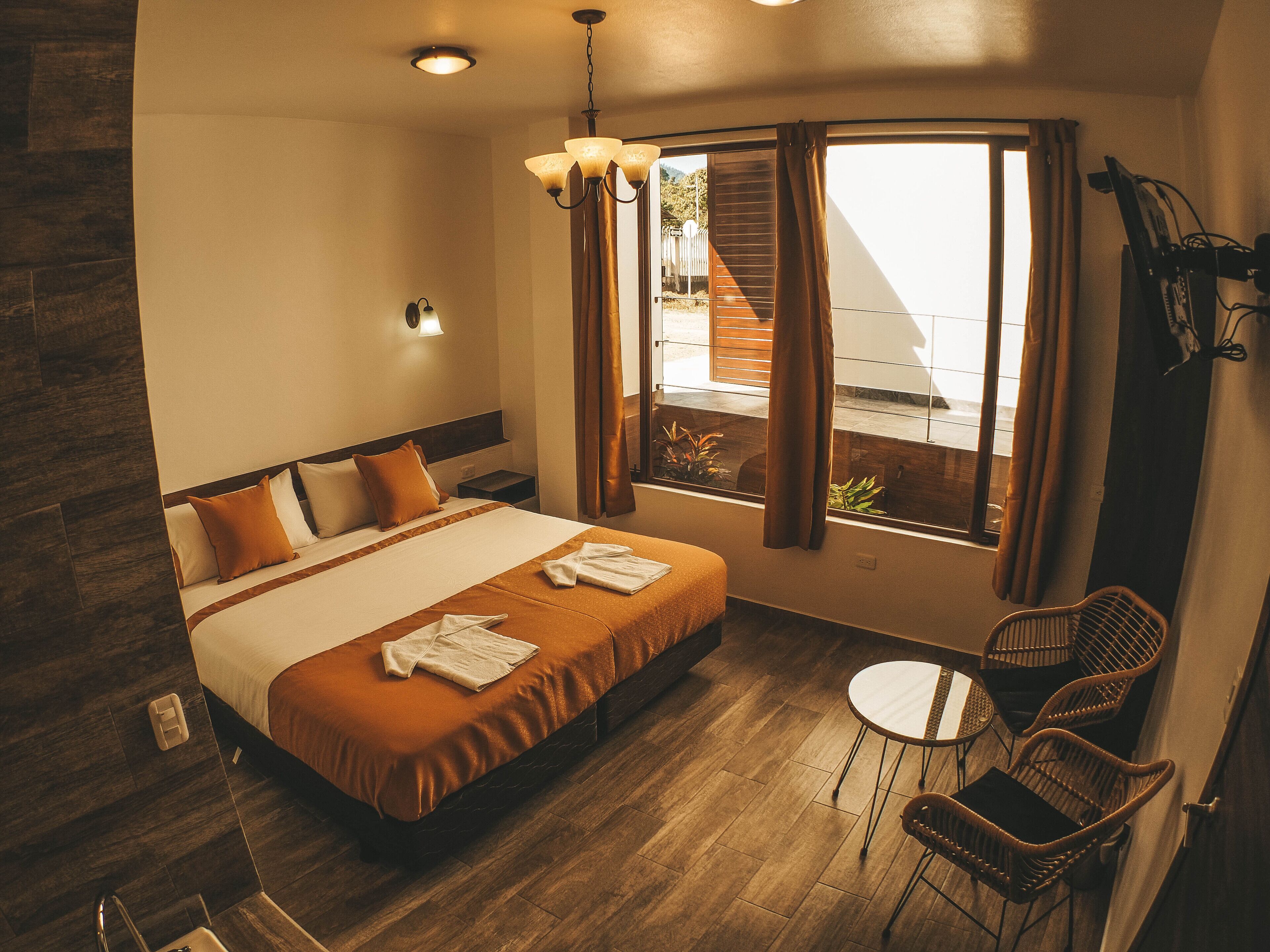 Habitación de hotel con cama, una mesita pequeña, sillas de mimbre, ventana con cortinas y vistas al exterior.
