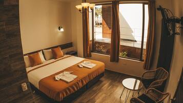 Habitación de hotel con cama, una mesita pequeña, sillas de mimbre, ventana con cortinas y vistas al exterior.