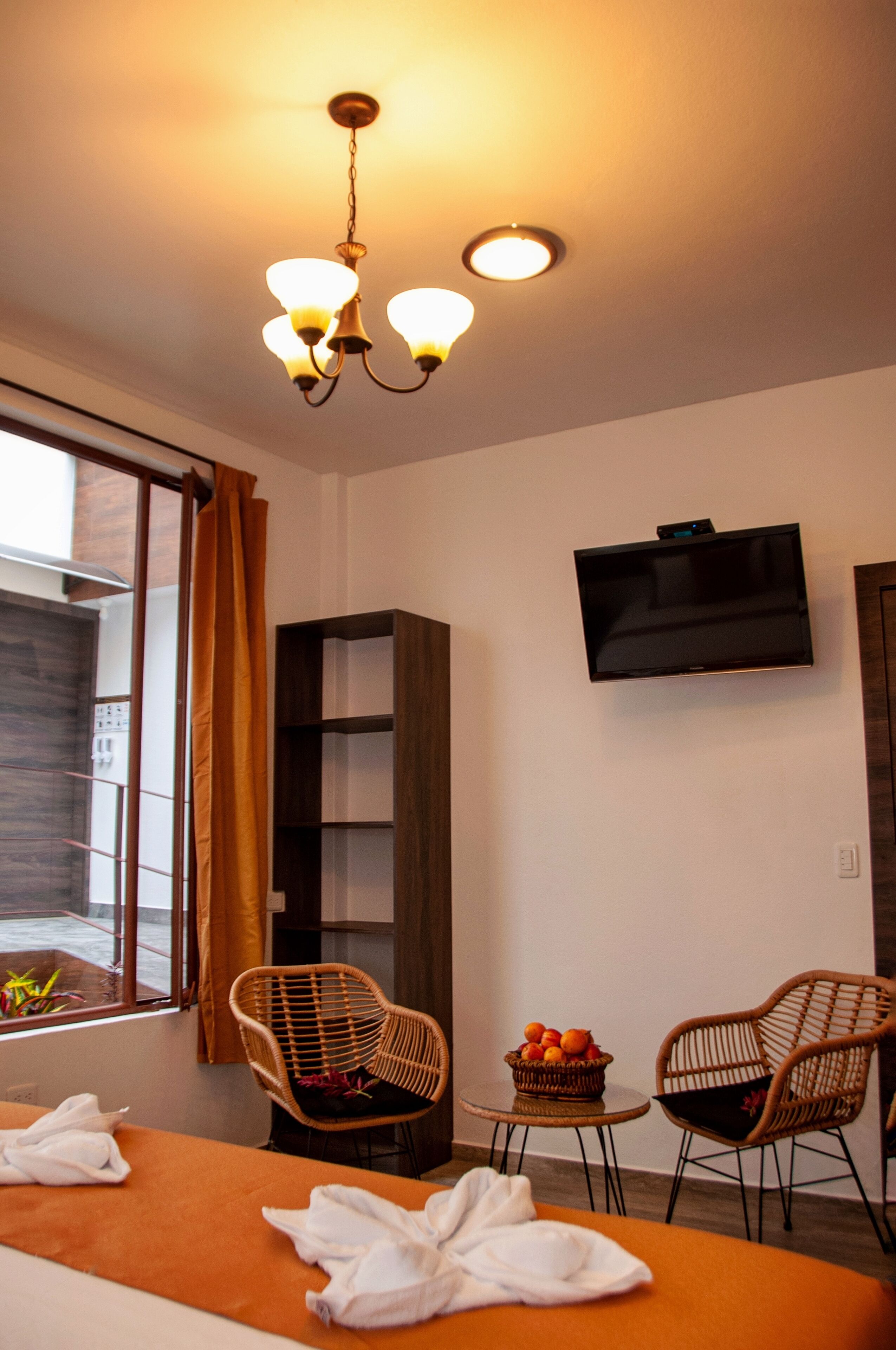 Une chambre d’hôtel avec un lit, deux fauteuils en osier, une petite table avec des fruits, une télévision et une fenêtre avec des rideaux.
