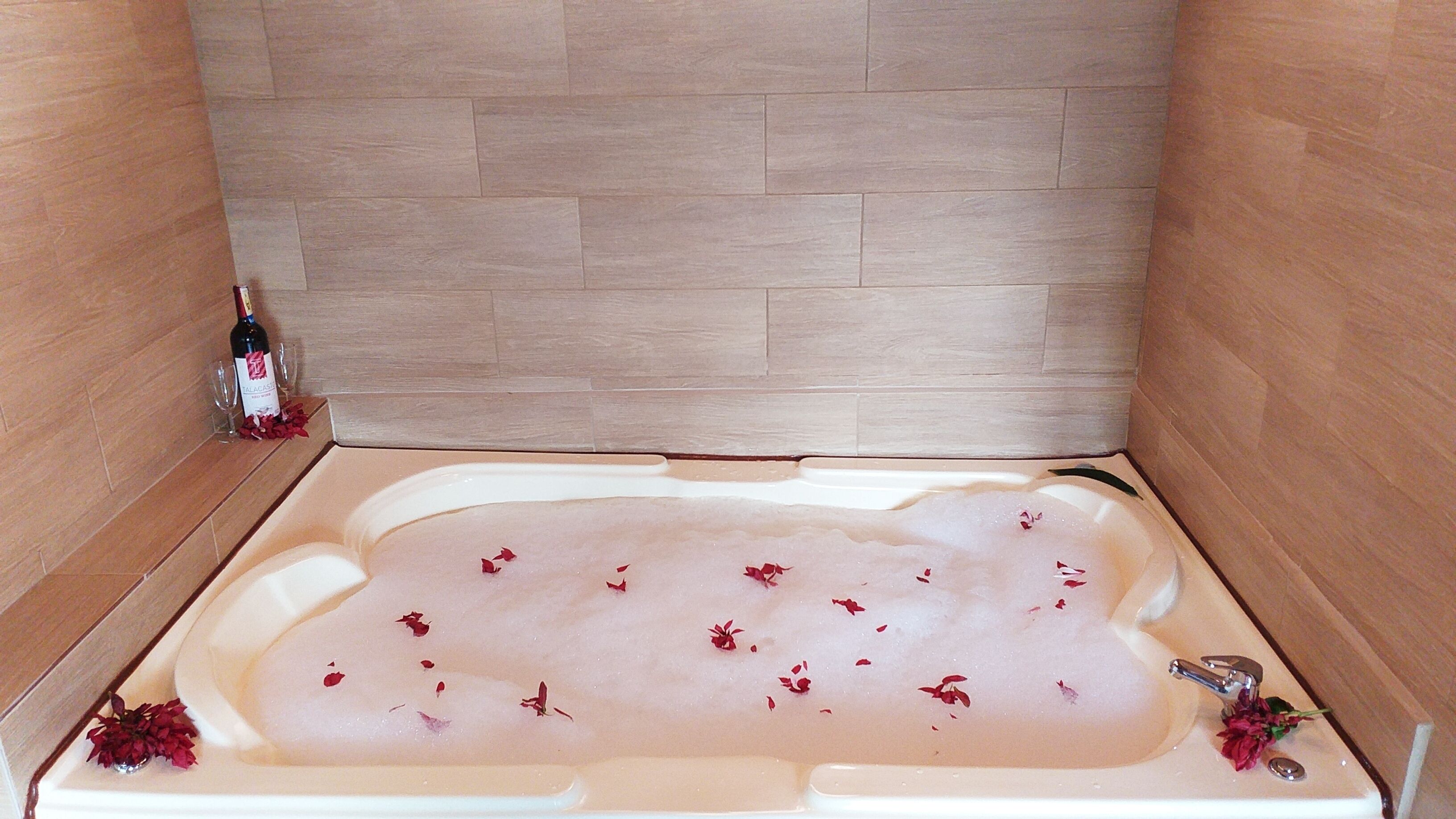 A bathtub filled with water and rose petals, with a wine bottle and glasses on the side.