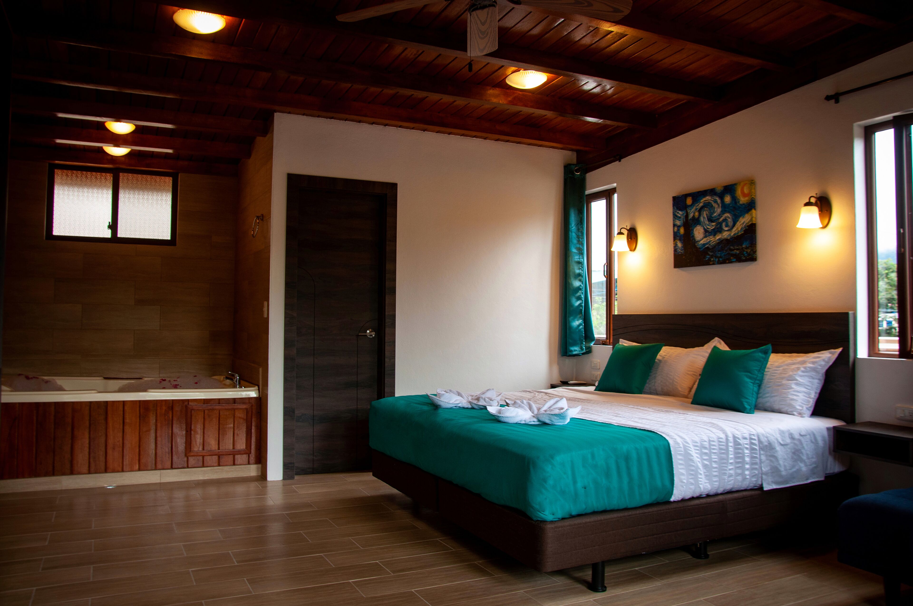 A hotel room with a wooden ceiling, a large bed with green and white bedding, a painting on the wall, and a window with curtains.