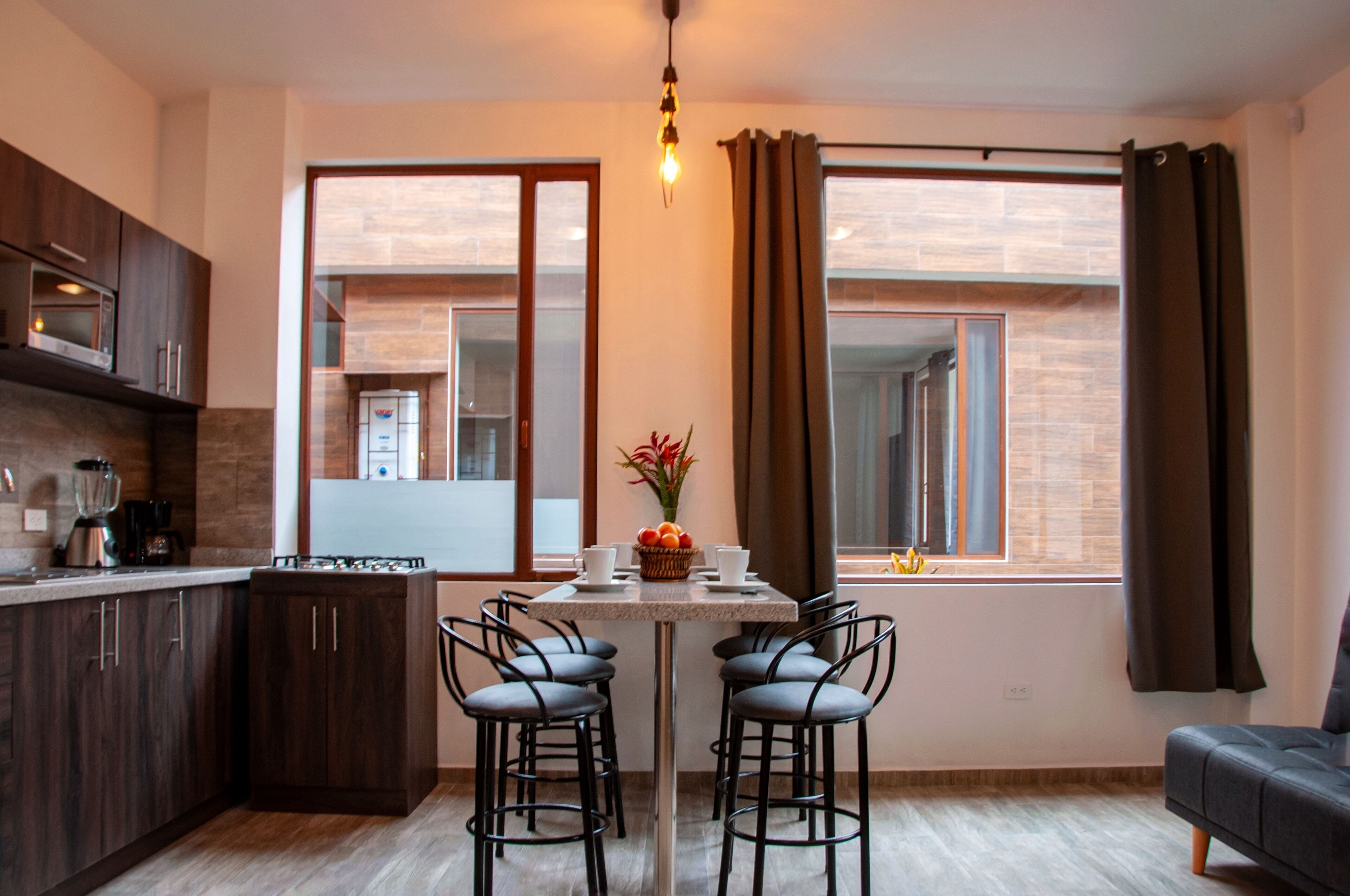 A modern kitchen with dark wood cabinets, a built-in oven, and a coffee machine. There is a dining area with a table and four chairs, and a sofa in the corner.