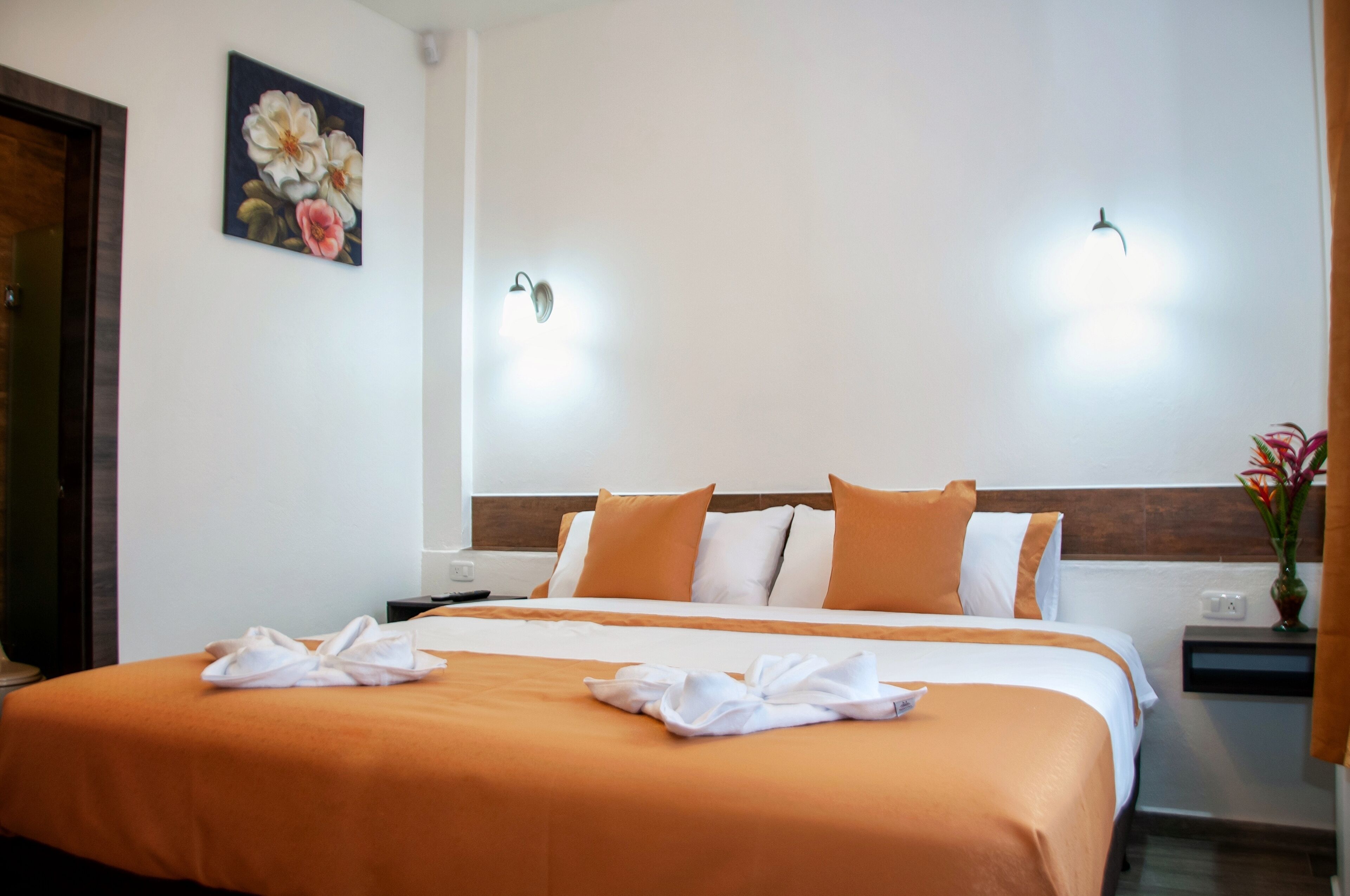 Une chambre d’hôtel avec un grand lit, deux lampes fixées au mur, un tableau et un vase rempli de fleurs.
