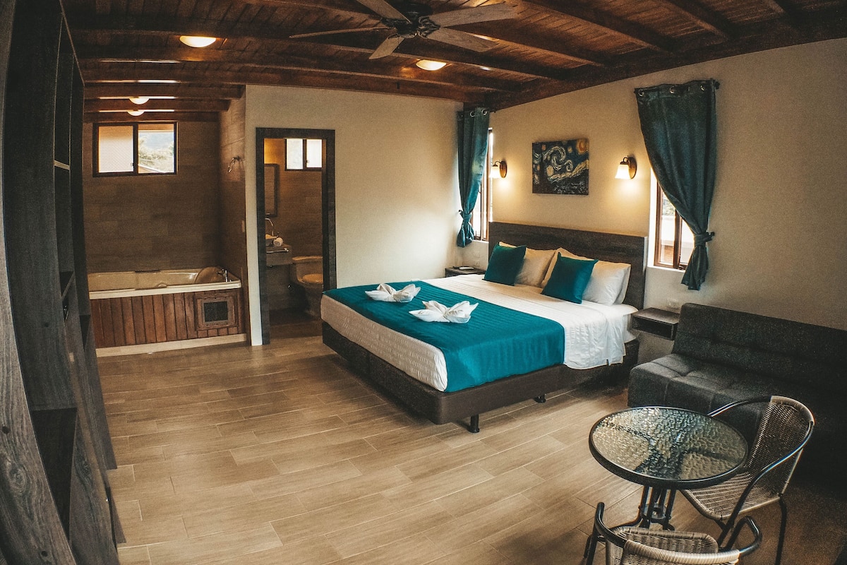 A hotel room with a wooden ceiling, a bed with a teal bedspread, a small round table, and a chair.