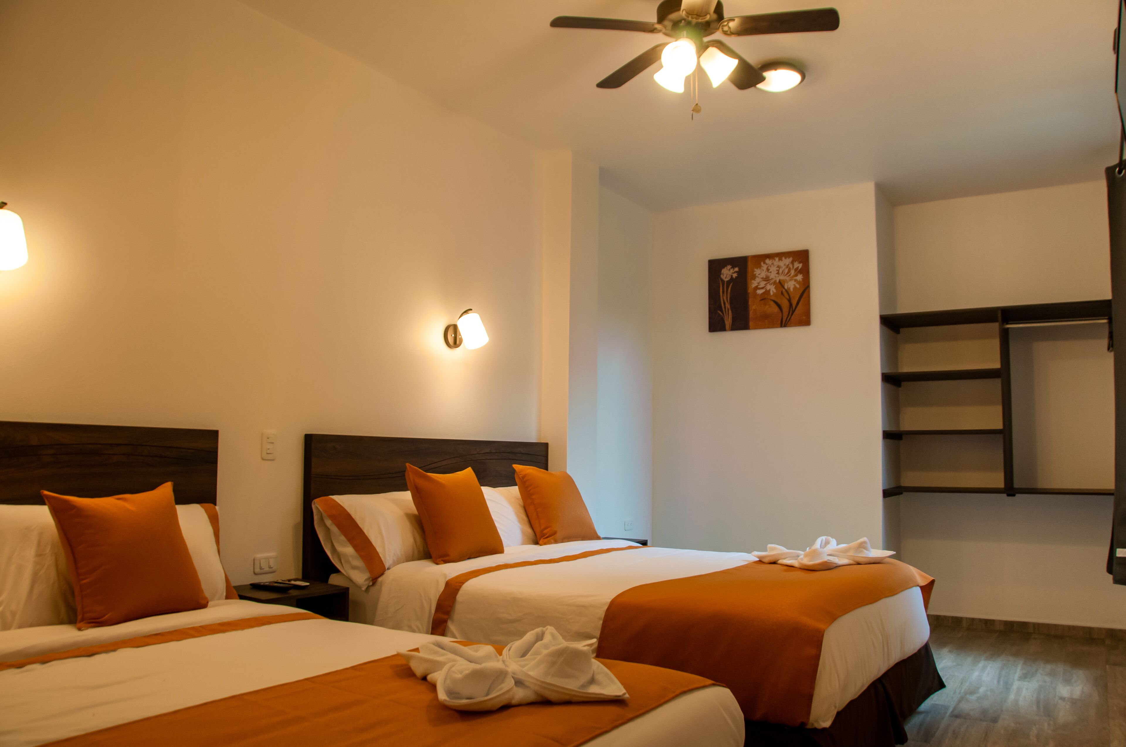 A hotel room with two beds, each with orange bedding and white pillows. There is a ceiling fan, wall-mounted lights, and a small painting on the wall.