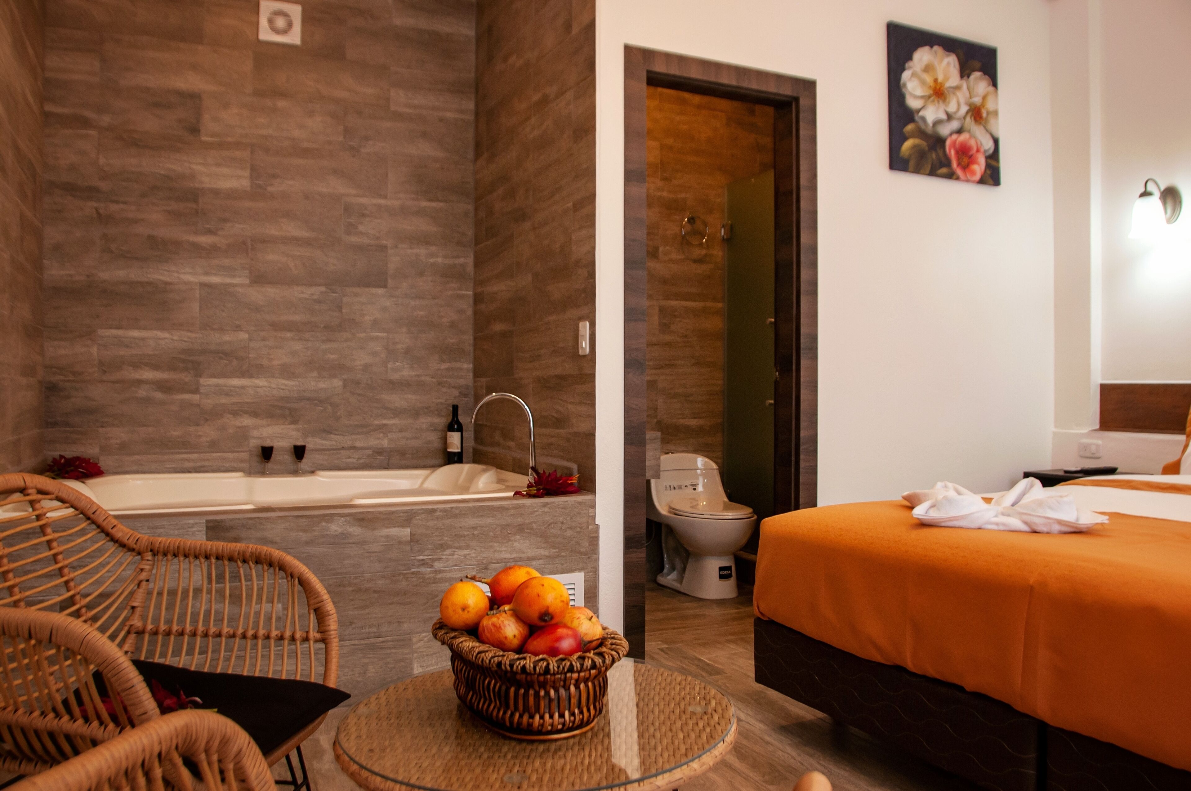 A hotel room with a bathtub, a toilet, a bed with an orange bedspread, wicker chairs, and a fruit basket on a round table.