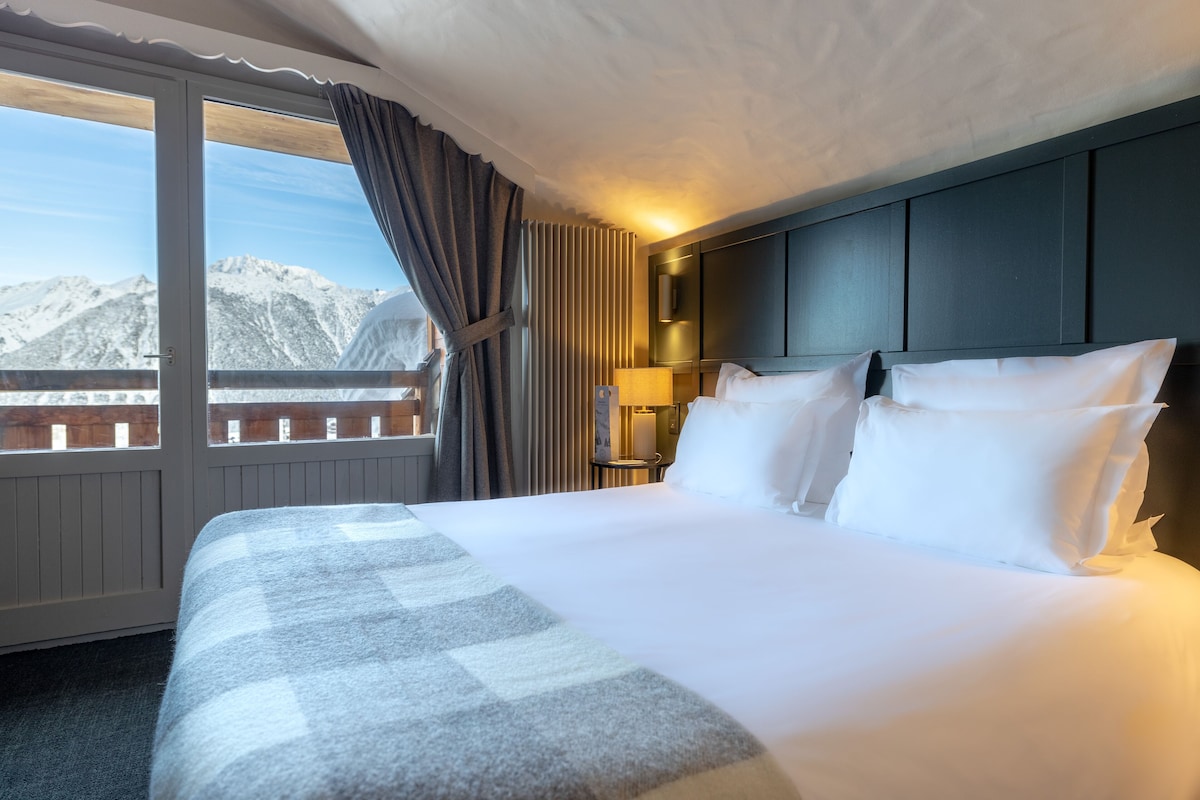A hotel room with a large bed, a view of mountains, a window with curtains, and a bedside table with a lamp.