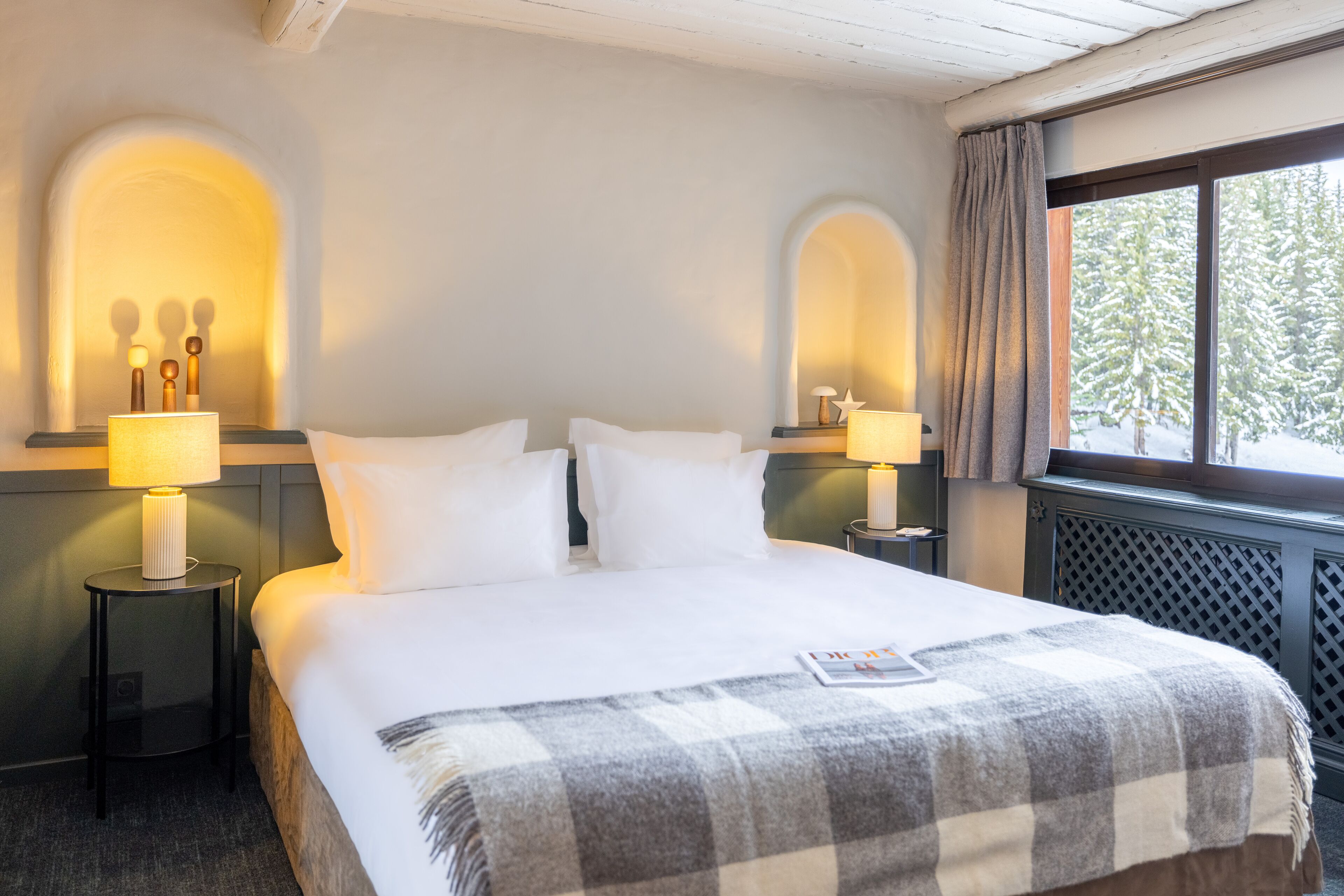 A hotel room with a large bed, two bedside lamps, a checkered throw blanket, and a window with a view of snow-covered trees.