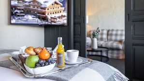 Bandeja de desayuno con cruasanes, fruta y jugo sobre una cama, con un televisor mostrando un paisaje nevado.