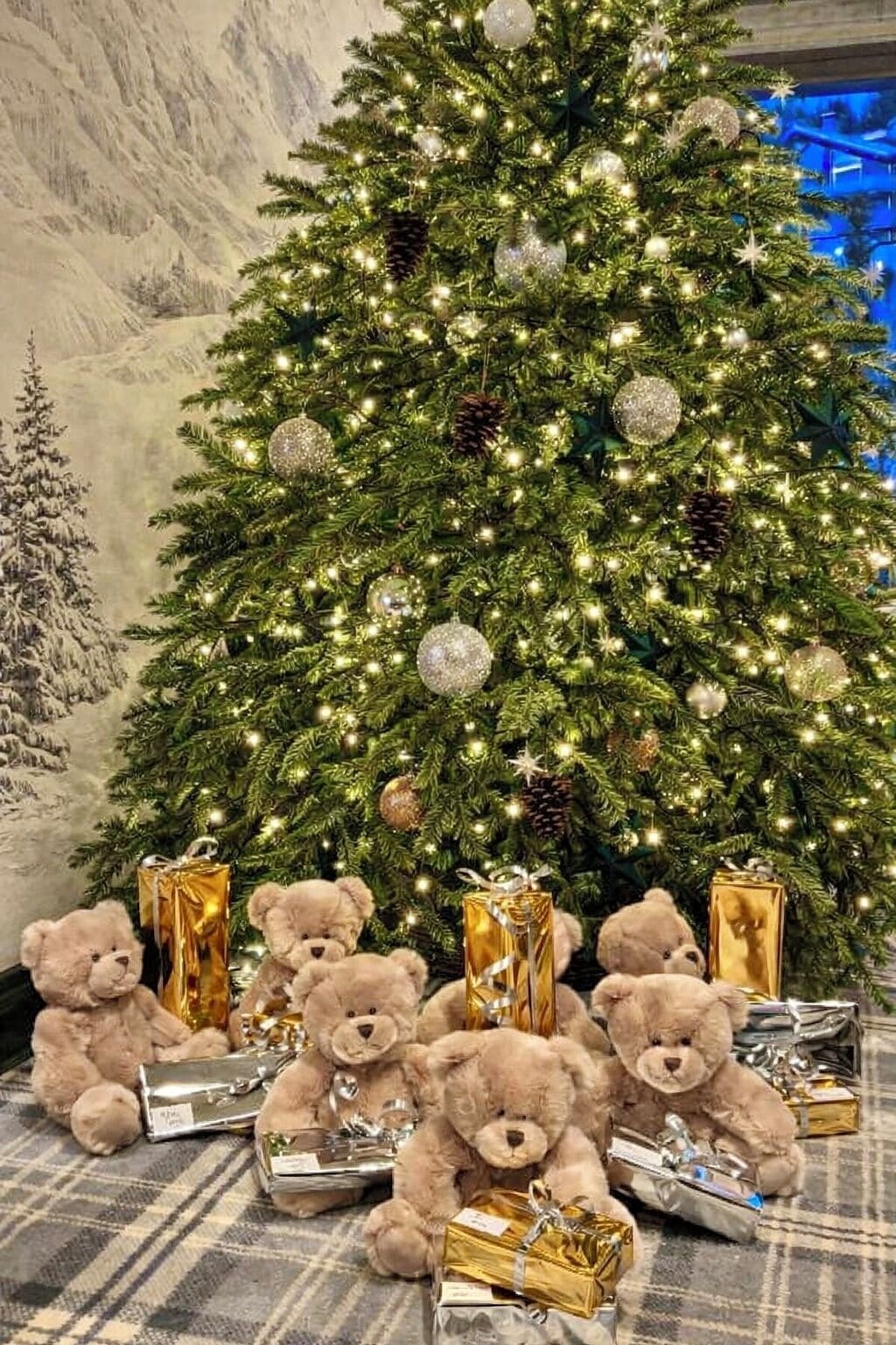 A Christmas tree with lights and ornaments, surrounded by teddy bears and wrapped gifts.