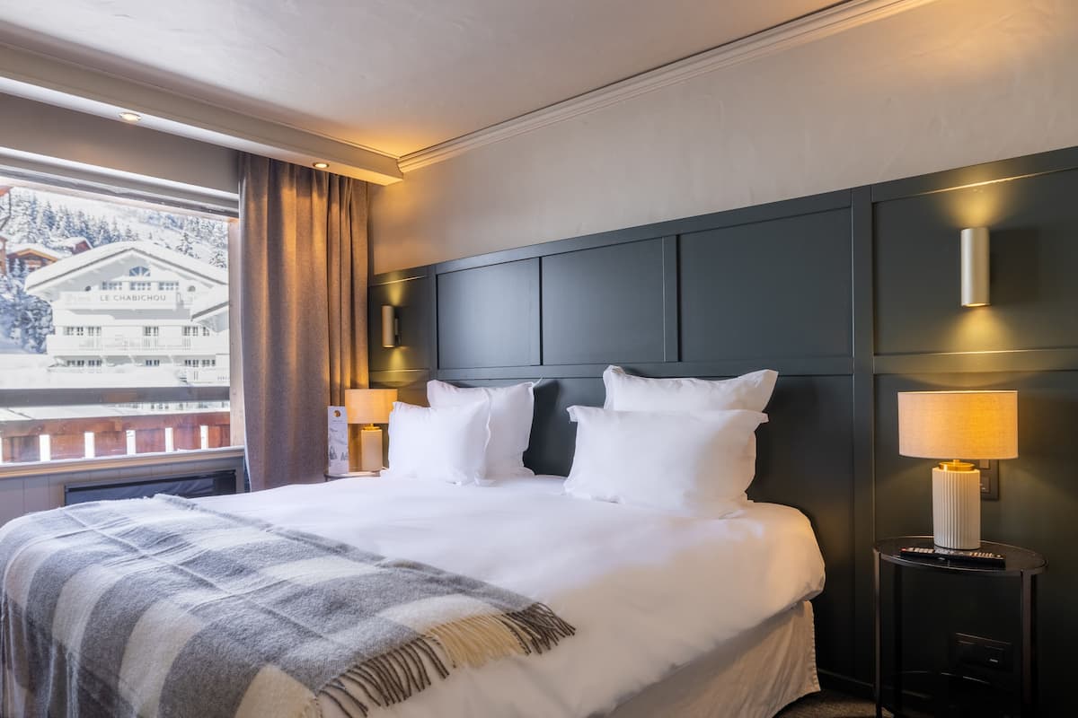 A hotel room with a large bed, two bedside tables with lamps, a view of a mountain and a building outside the window.