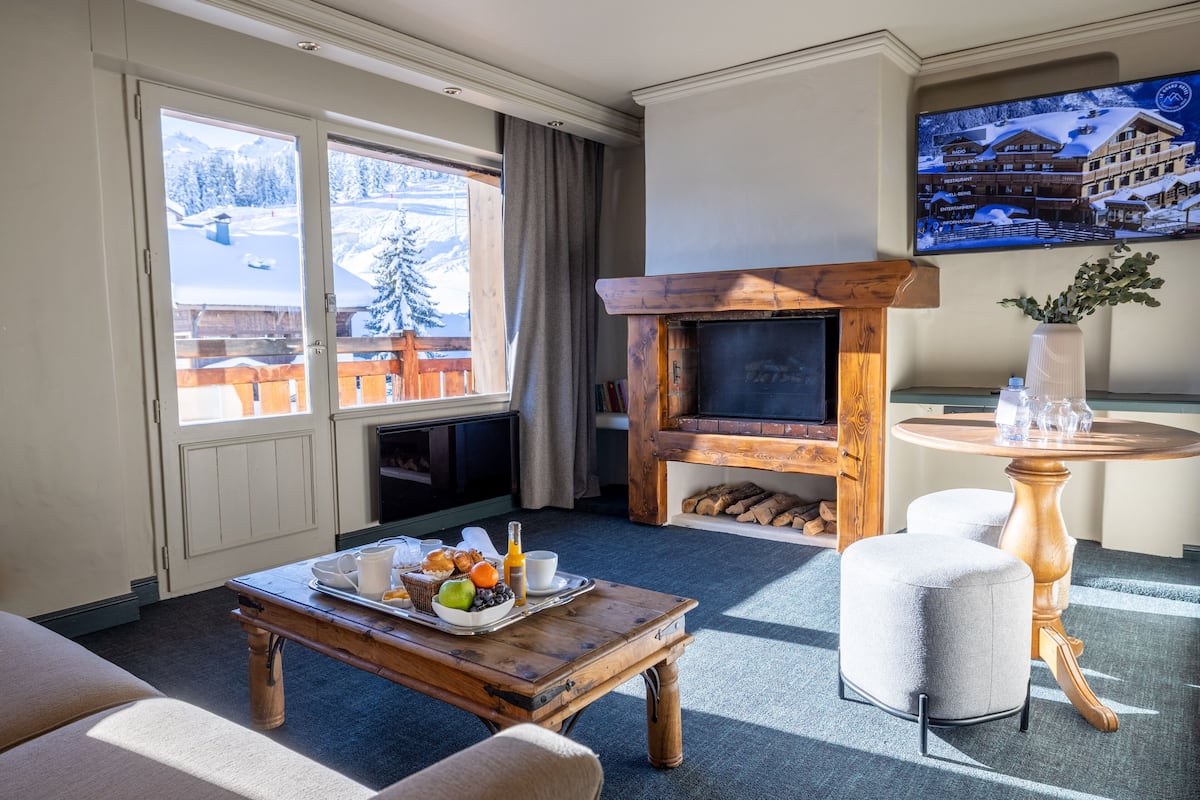 A cozy living room with a fireplace, a dining table with breakfast, and a large window with a snowy mountain view.