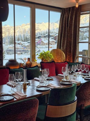 A dining area with a table set for six, large windows with a view of snow-covered mountains, and a plant on the windowsill.