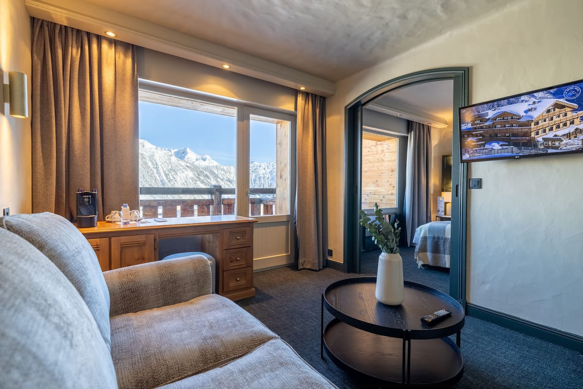A hotel room with a large window offering a mountain view, a cozy seating area, a small table with a vase, and a television on the wall.